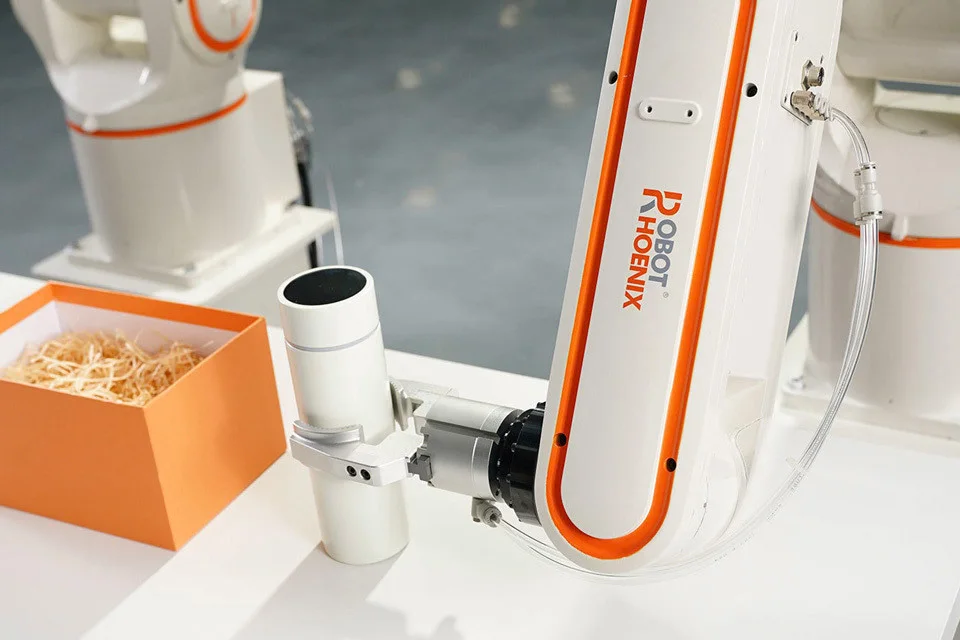 白い多関節ロボットのアームと、オレンジ色の箱が並ぶ作業台